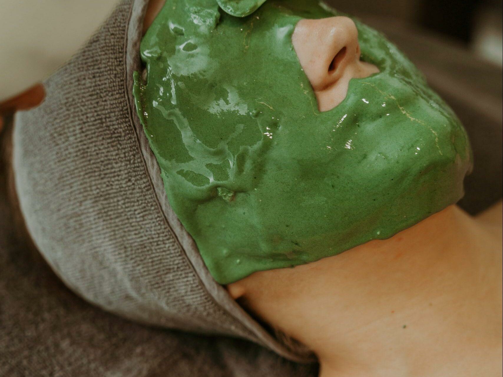 Esse-96 A person with their head wrapped in a gray towel is lying down and receiving a green facial mask. A hand is seen applying the mask with a spatula. The person's eyes are closed, and they appear to be relaxed. The background is blurred and neutral.