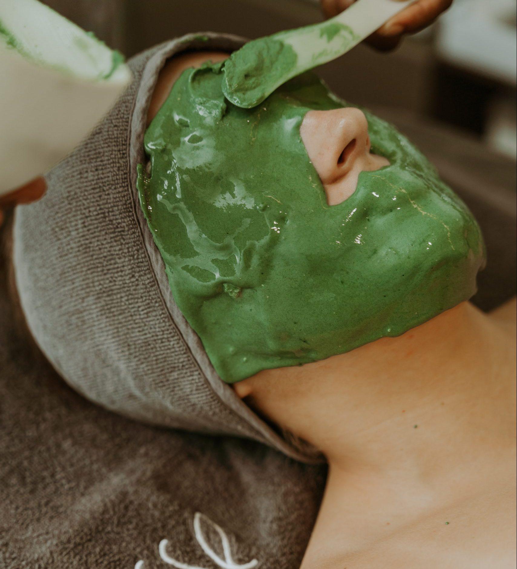 A person with their head wrapped in a gray towel is lying down and receiving a green facial mask. A hand is seen applying the mask with a spatula. The person's eyes are closed, and they appear to be relaxed. The background is blurred and neutral.