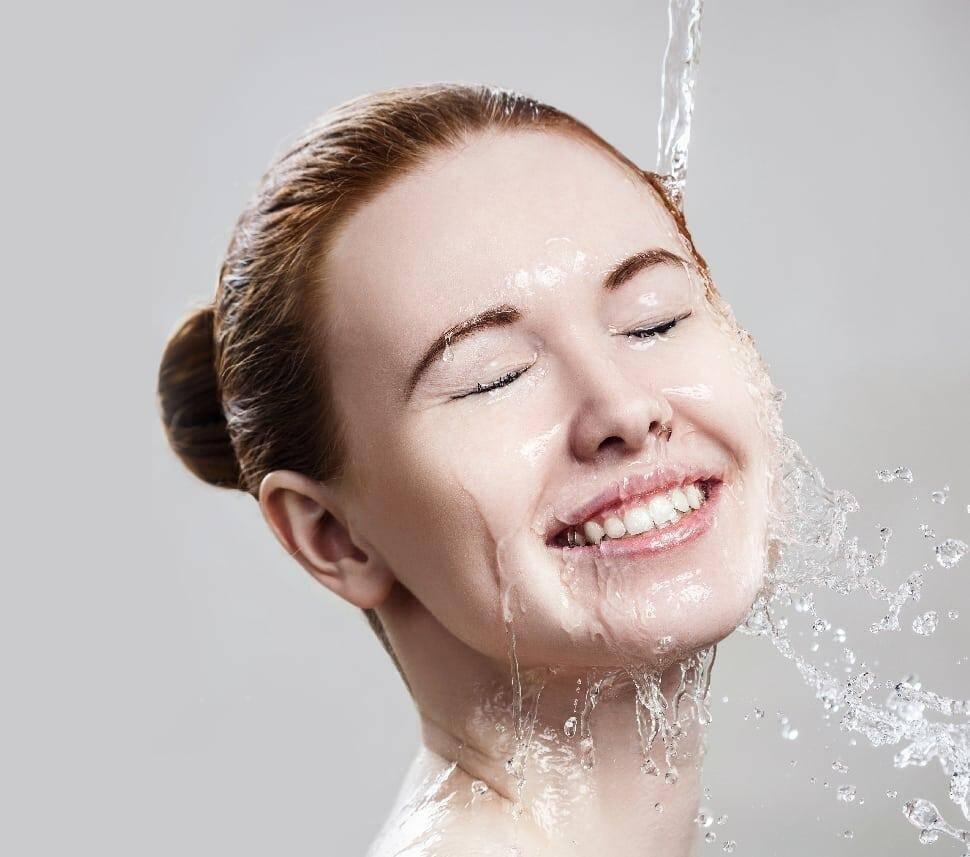 A person with their eyes closed and a joyful expression on their face tilts their head back under a stream of water, with droplets splashing around. Their hair is pulled back into a bun, and the background is a smooth, neutral shade.