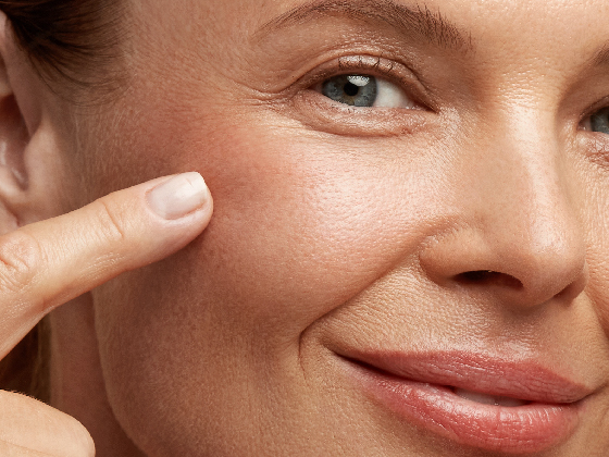 Close-up of a person gently touching their cheek with a finger. The skin appears smooth and moisturized. The person is smiling subtly and their hair and features are partially visible.