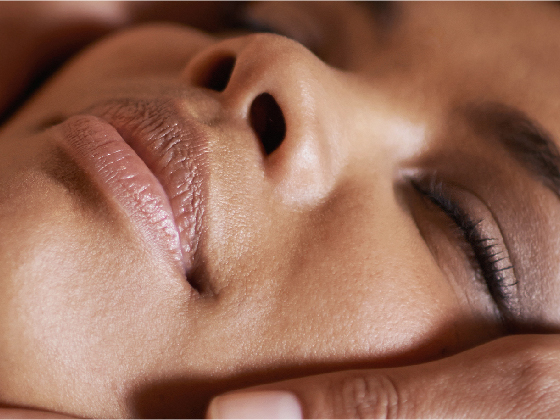 Close-up of a person's serene face with eyes closed, lips slightly parted, and a hand gently cradling the jaw. The lighting is soft, highlighting the smooth texture of the skin, suggesting relaxation and peace.