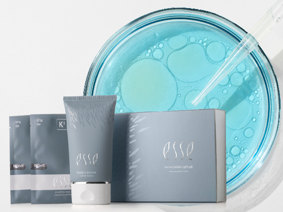 Skincare products including a tube and packets labeled "ESSE" are displayed in front of a glass dish filled with blue liquid. A dropper is resting on the dish, suggesting a scientific or cosmetic theme. The products and dish are on a white background.