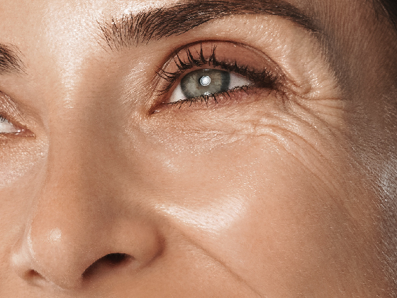 Close-up of a smiling person's eye and surrounding area, showing fine lines and a slightly furrowed brow. The skin has a natural glow, with light reflecting off the cheek and eyelid. Brown eyebrows and eyelashes frame the blue eye.