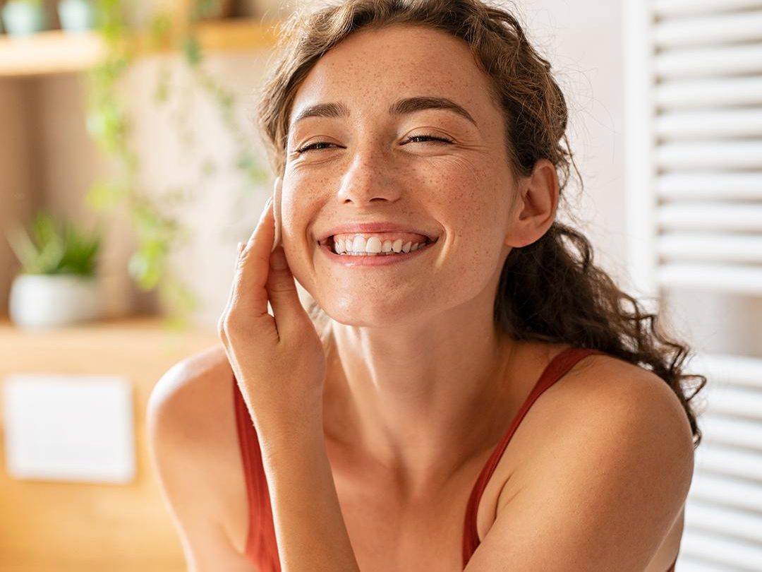 Sensitive Skin Guide – Image A woman with curly hair is smiling brightly and touching her face with one hand. She is wearing a sleeveless orange top. The background features a home setting with houseplants and wooden shelves, suggesting a relaxed and happy environment, perfect for those wondering, "What is sensitive skin?