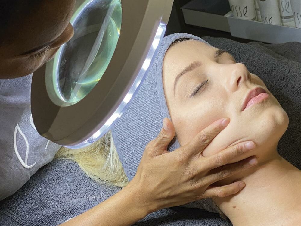 Apr 5 A person is receiving a facial treatment at a spa. Another individual is gently holding their face and examining it under a large magnifying lamp with built-in lights. Skincare products are visible in the background.