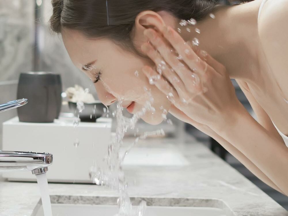 Cleansin The Esse Way – Image A woman with her hair tied back washes her face over a bathroom sink. Water splashes around her as she leans forward with her hands cupping the water from the running faucet. The bathroom has a modern design with a marble countertop and silver fixtures.