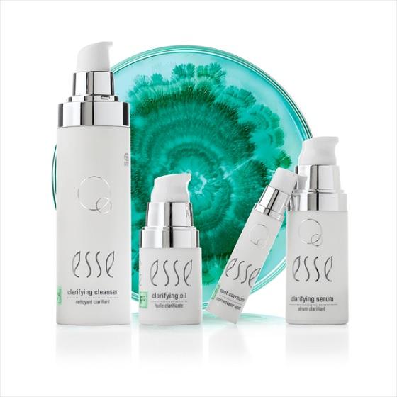 A product display featuring four white skincare bottles labeled "esse": a clarifying cleanser, clarifying oil, an eye product, and a clarifying serum. The items are arranged in front of a teal and white marbled background.