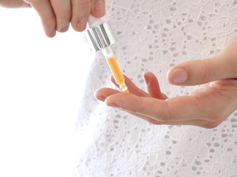 Close-up of a person applying a dropper of orange serum onto their finger. They are wearing a white, textured, lace-like top.