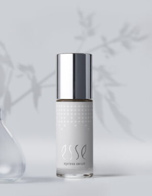 Esse Ageless Serum, capped in silver, sits on a light gray surface with a blurred plant shadow behind and part of a glass vase visible to the left.
