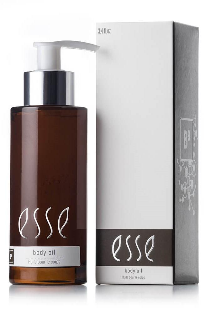 A bottle of Esse Body Oil, presented in a brown, transparent vessel equipped with a pump dispenser, positioned adjacent to its matching white and gray packaging box. The label on both the bottle and the box prominently displays the text "Esse Body Oil" and "Huile pour le corps.
