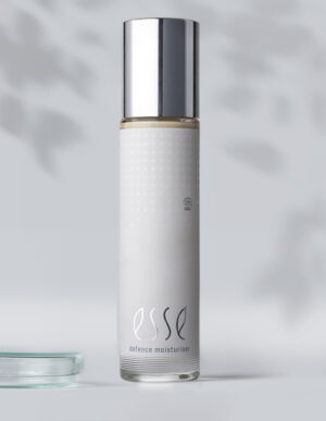 A sleek white bottle with a silver cap labeled "Esse Defence Moisturiser" stands upright on a light gray surface against a softly blurred background.