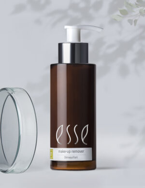 A brown bottle of Esse Make-up Remover with a white pump and a gray label stands against a light background with delicate shadowed leaves. The bottle's cap is also visible near the bottom left, and the product label includes the word "Démaquillant.