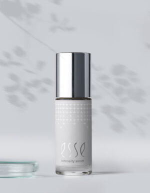 A bottle of Esse Intensity Serum with a silver cap stands on a light surface next to a partially visible glass dish, as soft leaf shadows fall across the minimal gray background.