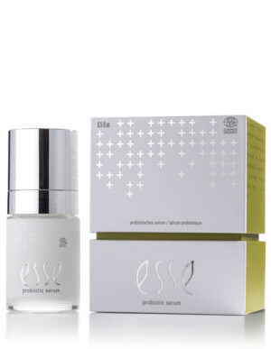 A white and silver bottle of Esse Probiotic Serum stands beside its matching box, both featuring silver details and a subtle cross pattern. The label reads "probiotic serum" and displays an organic certification logo.