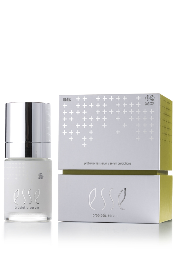 A white and silver bottle of Esse Probiotic Serum stands beside its matching box, both featuring silver details and a subtle cross pattern. The label reads "probiotic serum" and displays an organic certification logo.