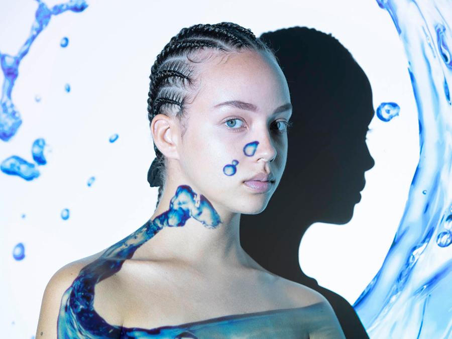 A young person with cornrow braids stares forward against a light background with artistic water splashes and droplets. Their shadow is prominently visible on the wall behind them. The overall color scheme has cool blue and white tones.