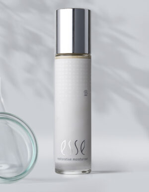 A tall, sleek bottle of Esse Restorative Moisturiser with a shiny silver cap stands against a light gray background featuring soft shadows, while a clear glass object is partly visible on the left.