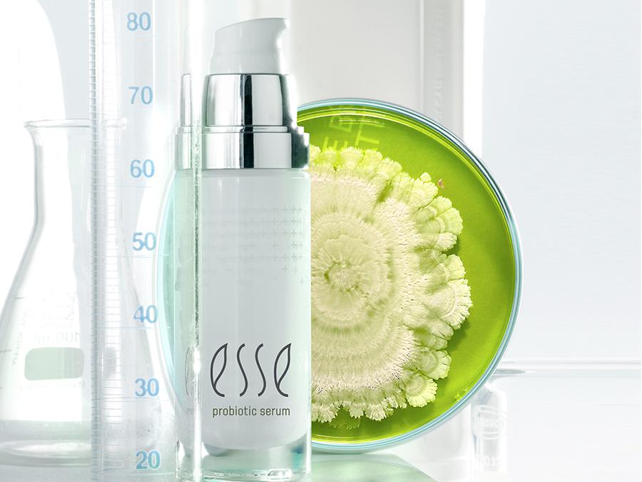 A bottle of Esse Probiotic Serum is positioned against a backdrop of laboratory equipment including a beaker, measuring cylinder, and a Petri dish containing a green, flower-like culture. The scene evokes a scientific and skincare-themed setting.