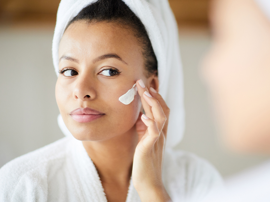 A woman with a towel wrapped around her head and wearing a bathrobe applies a cream to her cheek. She is looking in the mirror while gently using her fingers to rub in the product. The setting appears to be a bathroom.