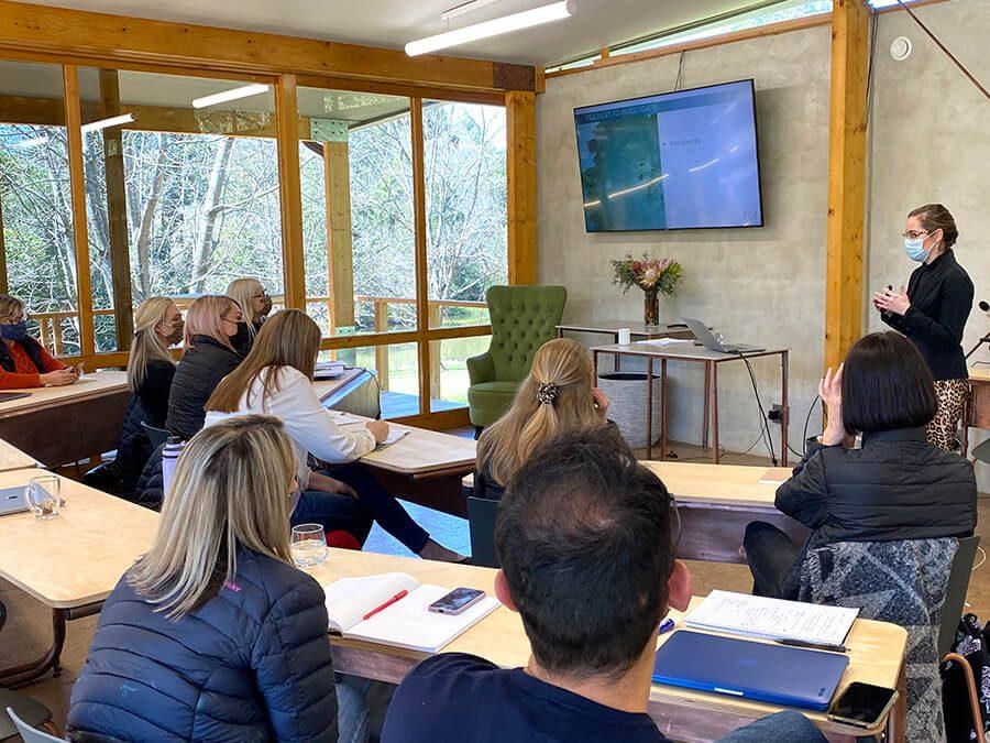 A group of people sit in a classroom facing a woman in a mask who is giving a presentation. The classroom has large windows with a view of trees. The presenter stands near a green armchair, and a large screen displays content beside her.