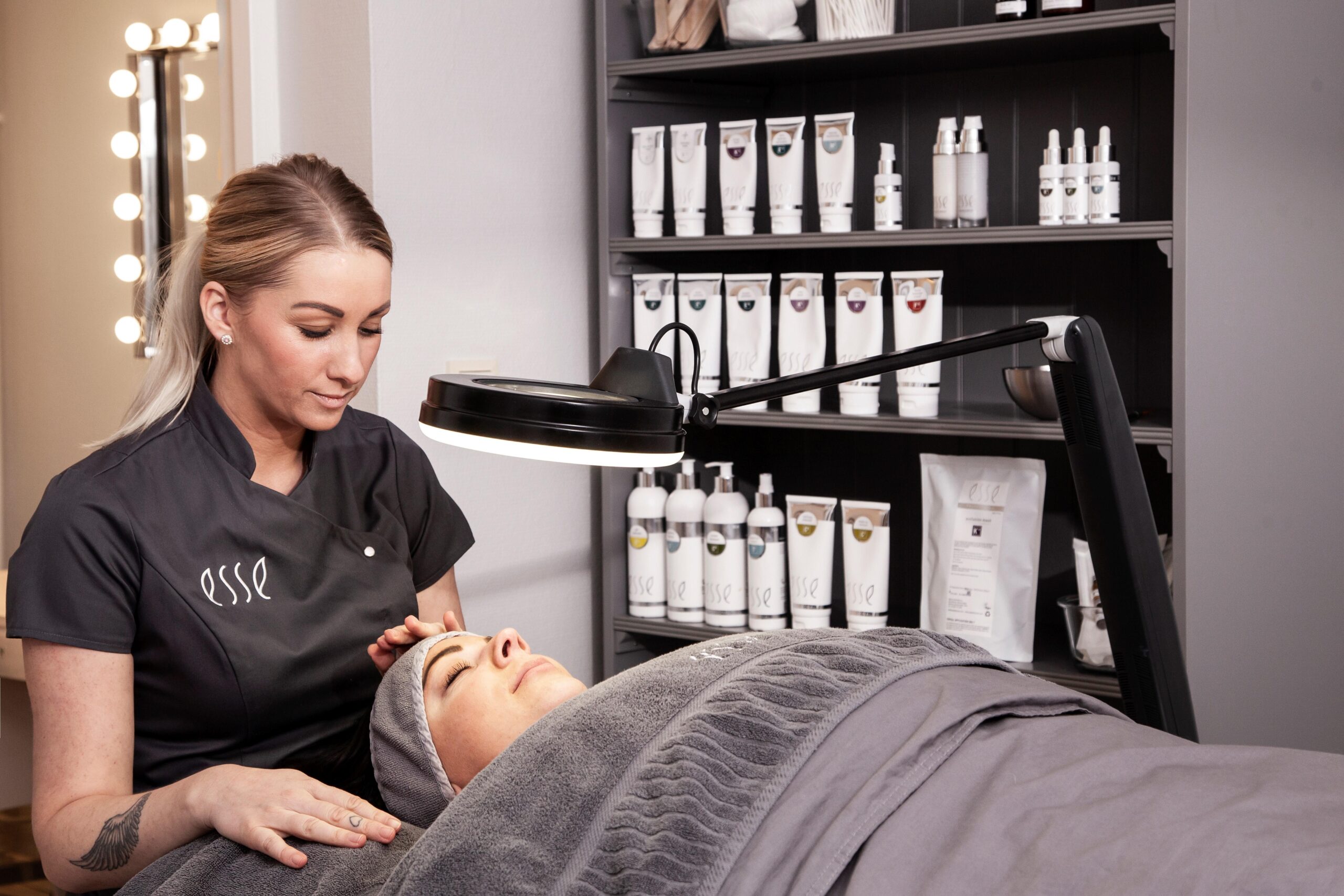 An esthetician wearing a gray uniform is giving a facial treatment to a client lying on a treatment table covered with gray blankets. The room has shelves stocked with various skincare products and a magnifying lamp positioned over the client's face.