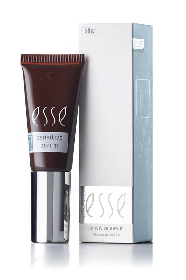 A sleek tube of Esse Sensitive Serum, in a sophisticated dark color, features a metallic silver cap and stands beside its light grey packaging box adorned with subtle decorative patterns. Both the box and the tube display the product name and a volume of 0.5 fl oz.
