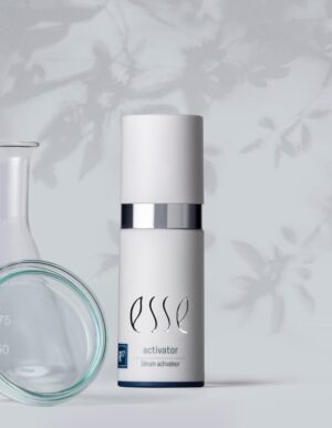 A white bottle labeled "Esse Activator Sérum Activateur" stands upright with a silver cap. It is placed beside clear laboratory glassware, including a beaker and a petri dish. The background has a light gray hue with a subtle floral pattern.