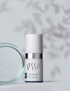A white bottle labeled "Esse post-care oil" is placed against a light, neutral backdrop with soft shadows of foliage. The bottle has a minimalist design and a silver band near the top. A clear glass lid or container lies beside it, adding to the clean aesthetic.