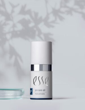 A bottle of Esse pre-care oil with a white cap and silver accent stands against a light grey background. Shadows of leaves are cast on the wall behind it, creating a natural, serene atmosphere. To the left of the bottle is a small glass dish.
