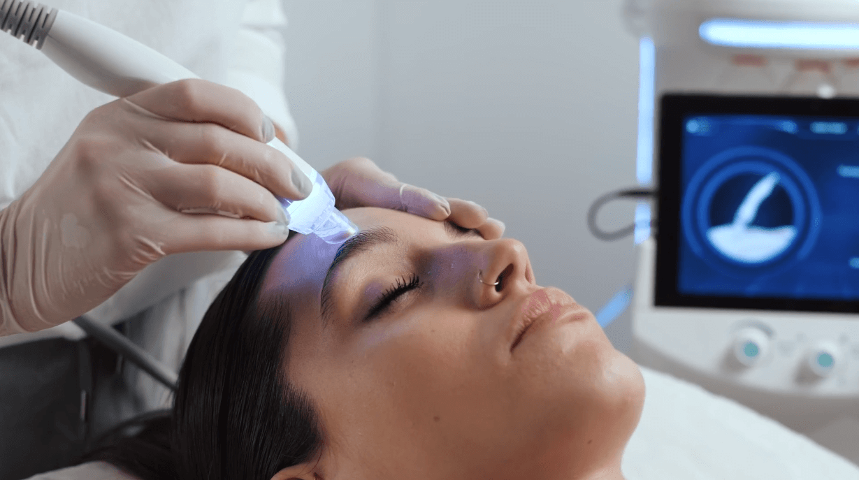 A person lies with their eyes closed while a gloved professional uses the Esse Activator, a handheld device, to perform a facial treatment on their forehead. A monitor displaying information is visible in the background. The setting appears to be a clinic or spa.