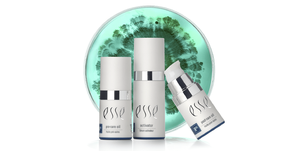Three Esse skincare product bottles are displayed in front of a dish with a green and teal gradient pattern. The products include pre-serum oil, activator, and a third bottle with a pump dispenser. All bottles are white with silver accents and branded "Esse.
