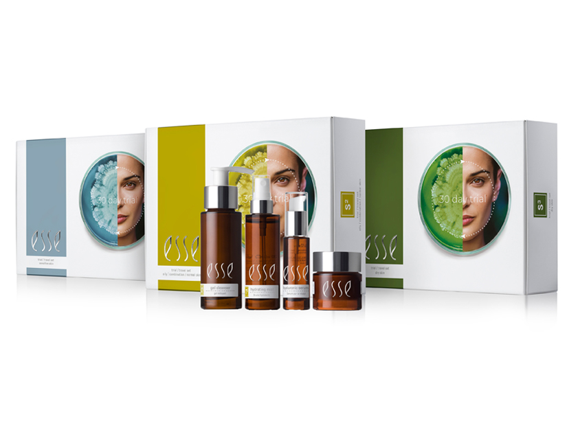 A display of skincare products from the brand Esse, featuring various bottles and containers in front of three boxes with images of a face on each. The packaging colors are predominantly white, blue, yellow, and green.