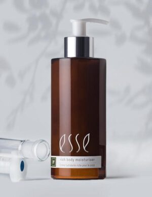 A brown bottle of Esse rich body moisturiser with a pump dispenser. The label has the brand name and product description in white text. The background is softly lit and includes blurred shadowed foliage, accompanied by a clear plastic cylindrical item near the bottle.