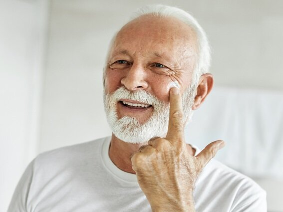 1039786d-772f-43c8-aeaf-610bf2d3de3b An elderly man with white hair and a beard smiles as he applies a cream to his cheek using his finger. He is wearing a white shirt and appears to be in a well-lit room, possibly a bathroom or bedroom.