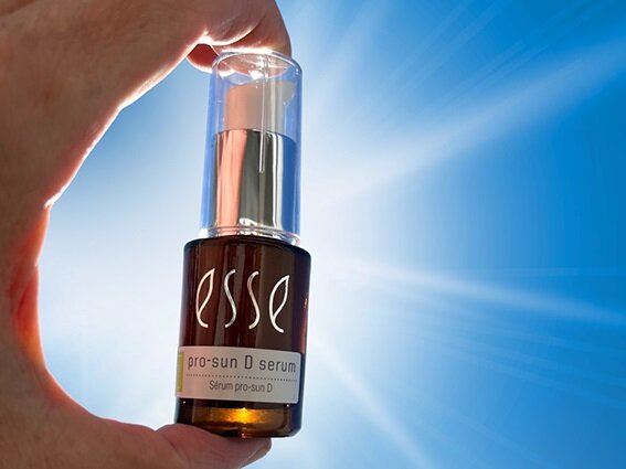 A hand is holding a small amber bottle of Esse Pro-Sun D Facial Serum against a bright, sunlit sky. The bottle has a silver pump dispenser and a white label with text. Sun rays radiate in the background, highlighting the product.
