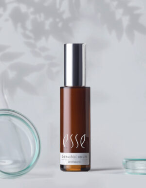 A brown glass bottle labeled "Esse Bakuchiol Serum" with a silver cap stands centered on a light grey surface. To the left, a lid enhances visual balance, while another lid lies flat in the bottom right corner. Shadows of branches add elegance to the background.