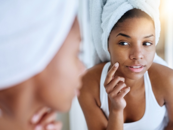 A woman with a towel wrapped around her hair looks at herself in the mirror, gently touching her cheek with one hand. She appears to be inspecting her skin. The setting suggests a bathroom, and she is wearing a white tank top.