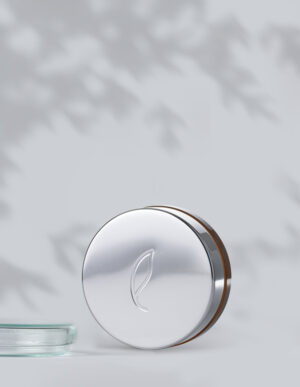 A round, silver tin labeled Esse Concealer with an embossed leaf design on the lid stands against a light gray background. The tin is partially open, revealing a hint of the concealer inside. Shadows of leaves create a subtle pattern on the wall in the background.