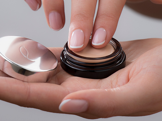 Close-up of a person's hands, with one hand holding a small jar of cream and the other dipping a finger into the cream. The person has manicured nails with a light pink polish and white tips. The jar has a shiny metal lid resting on the palm.