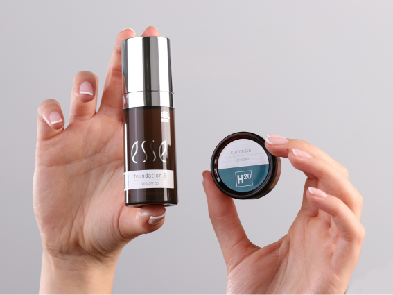 Two hands hold beauty products against a plain background. The left hand displays a dark bottle labeled "foundation 3". The right hand presents a round container labeled "concealer H2O". Both hands have well-manicured nails with a light polish.
