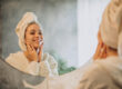 A person wrapped in a towel smiles at their reflection in a bathroom mirror while gently touching their face. The background features a leafy plant and soft, natural lighting.