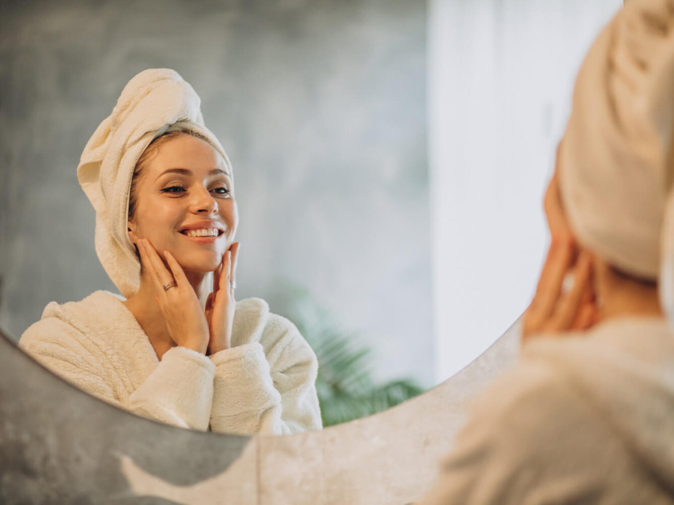 24618 A person wrapped in a towel smiles at their reflection in a bathroom mirror while gently touching their face. The background features a leafy plant and soft, natural lighting.