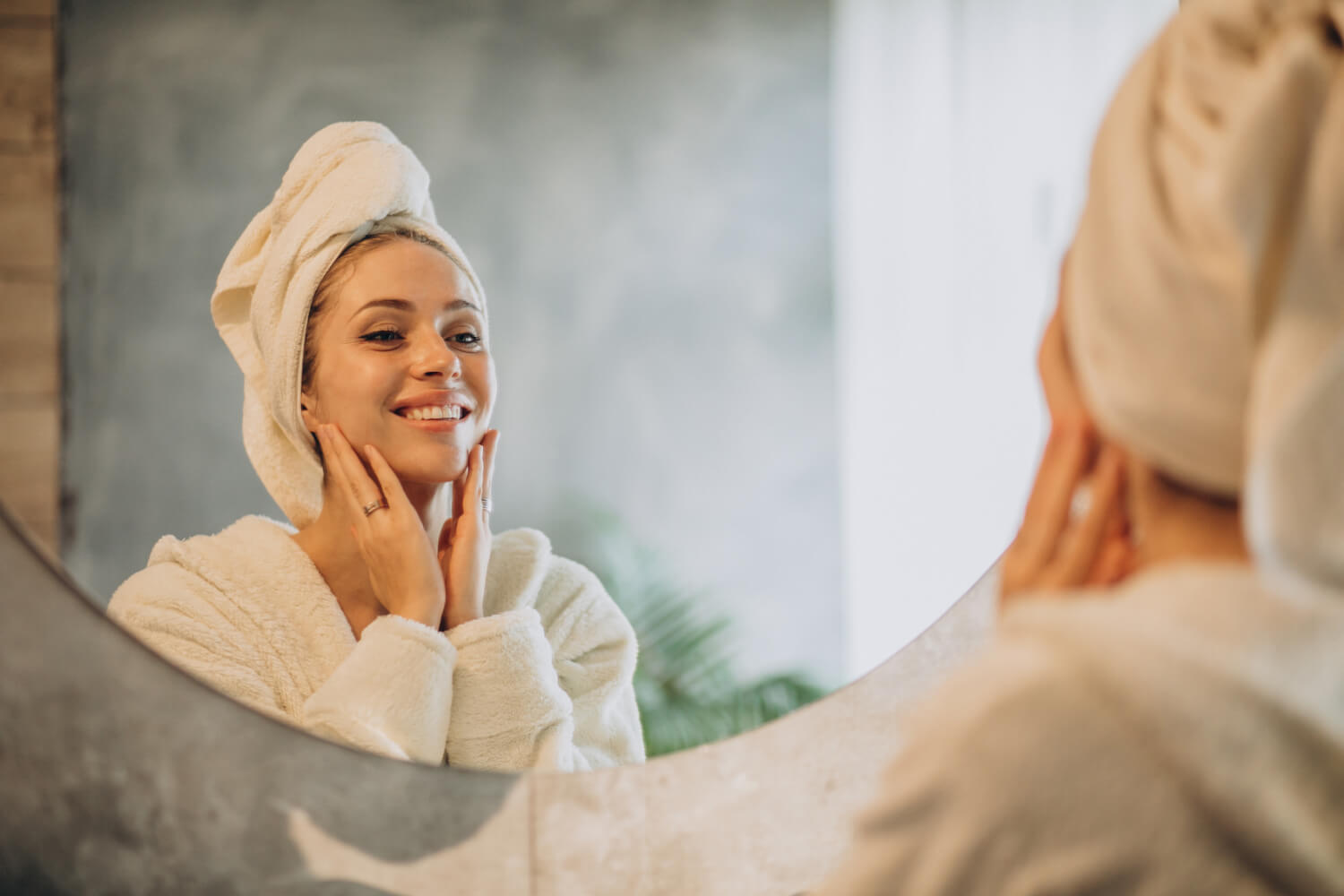 A person wrapped in a towel smiles at their reflection in a bathroom mirror while gently touching their face. The background features a leafy plant and soft, natural lighting.