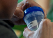 A person holds a transparent plastic container with a blue lid labeled "Propionic Acid." The focus is on the container and the person's hands, with a blurred background containing greenery.