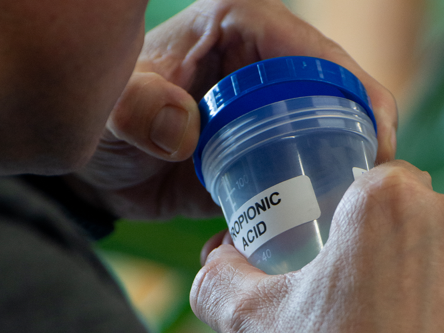 Blog Propionic Acid A person holds a transparent plastic container with a blue lid labeled "Propionic Acid." The focus is on the container and the person's hands, with a blurred background containing greenery.