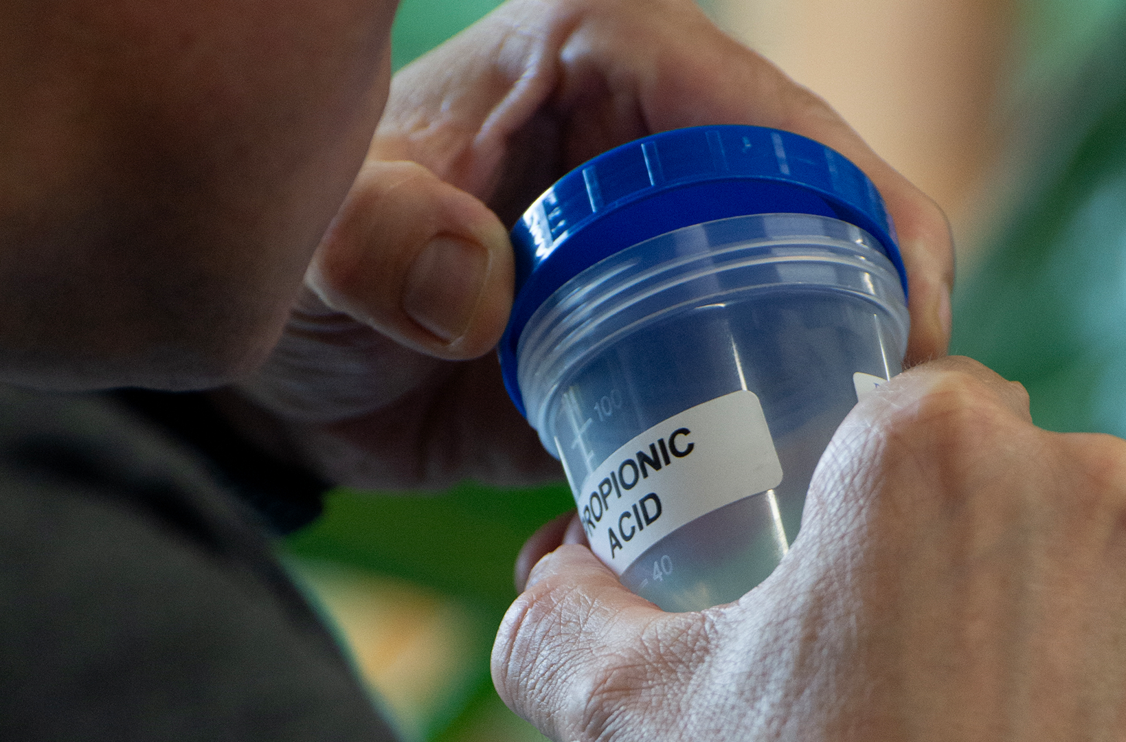 A person holds a transparent plastic container with a blue lid labeled "Propionic Acid." The focus is on the container and the person's hands, with a blurred background containing greenery.