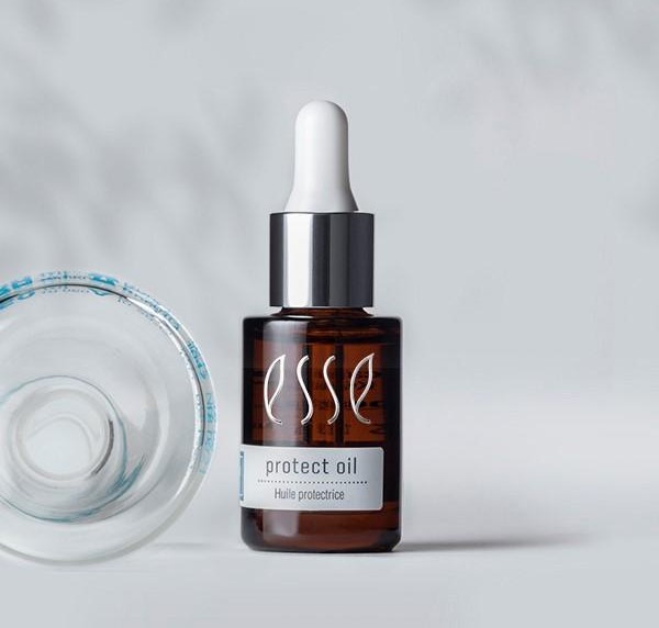 A brown dropper bottle labeled "Esse Protect Oil" stands upright with a silver cap and white dropper. Next to it, a clear, open lid lies on its side. The background is a soft gray with a subtle shadow pattern.