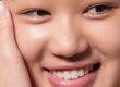 Close-up of a person smiling, showing smooth, glowing skin and white teeth. One hand gently touches their cheek. The image highlights healthy skin and a happy expression.