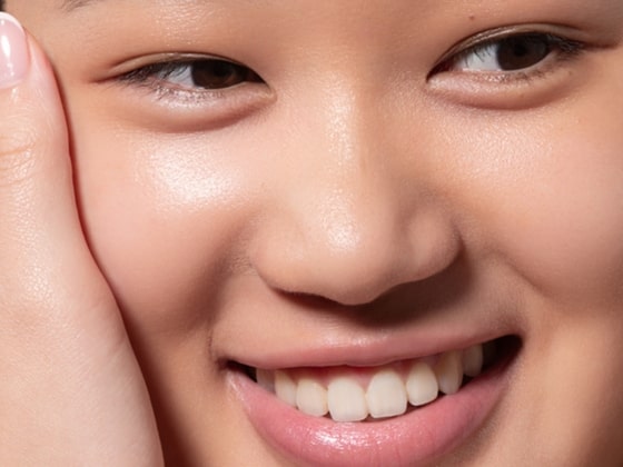 Close-up of a person smiling, showing smooth, glowing skin and white teeth. One hand gently touches their cheek. The image highlights healthy skin and a happy expression.
