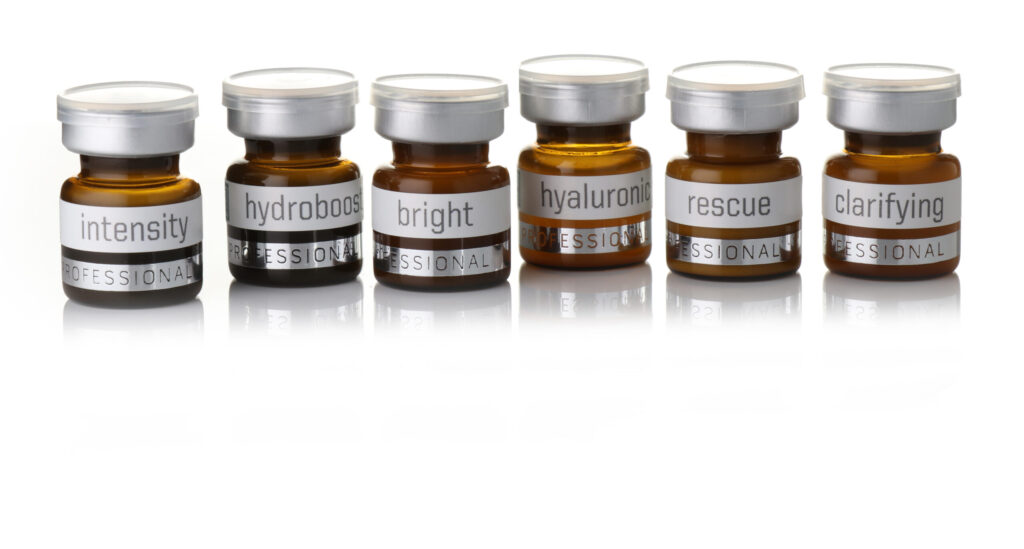 Six small brown skincare bottles with silver lids are lined up, each labeled: intensity, hydroboost, bright, hyaluronic, rescue, and clarifying. The bottles are reflected on the white surface below them.